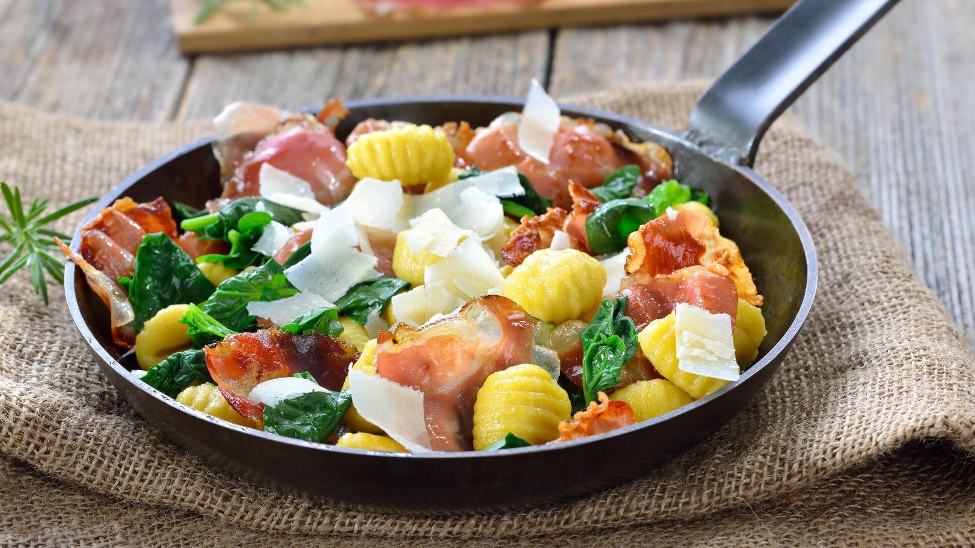 Gnocchi, Spinat, Speck und Parmesan in einer gusseisernen Pfanne. Diese steht auf einem Leinentuch.