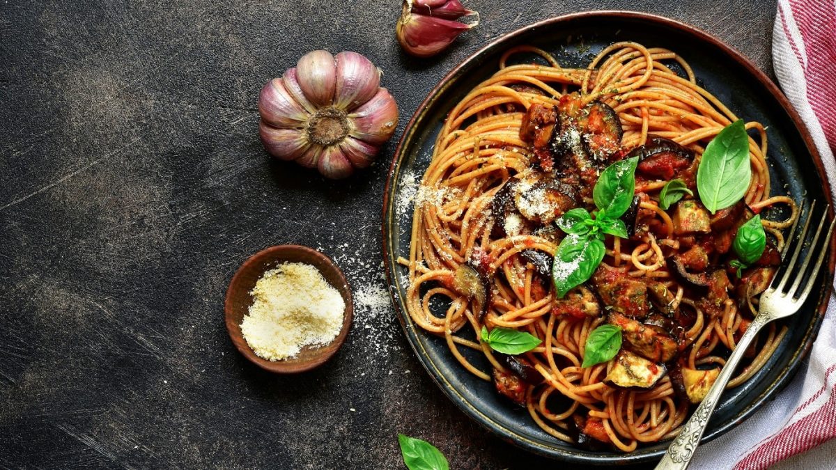 Spaghetti mit italienischer Norma-Sauce auf dunklem Teller. Das Ganze steht auf dunklem Untergrund und ist umgeben von lila Knoblauch und einem Schälchen Hartkäse.
