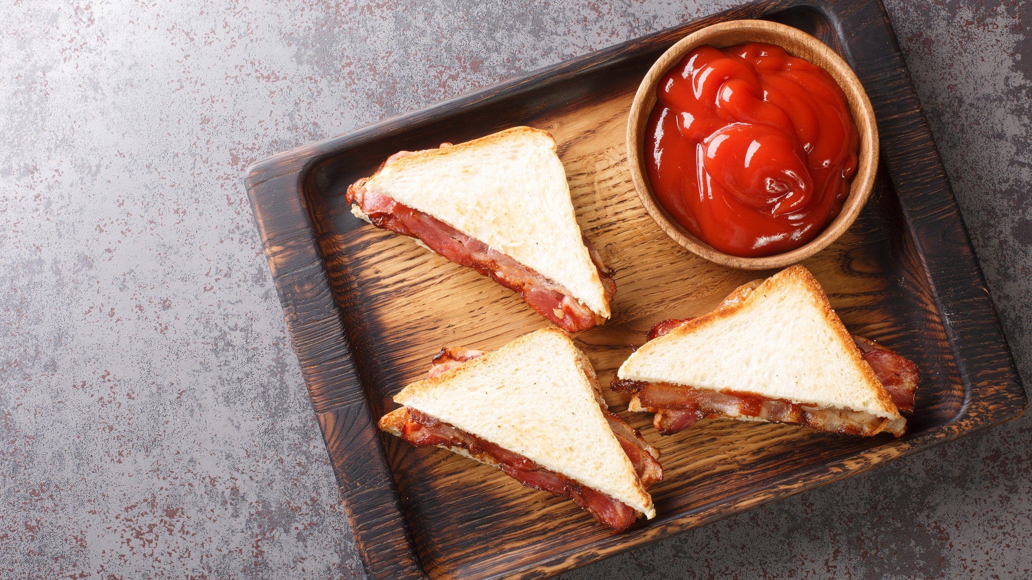 Bacon Butty Sandwich auf einem hölzernen Tablett. Daneben eine Schale mit Ketchup.