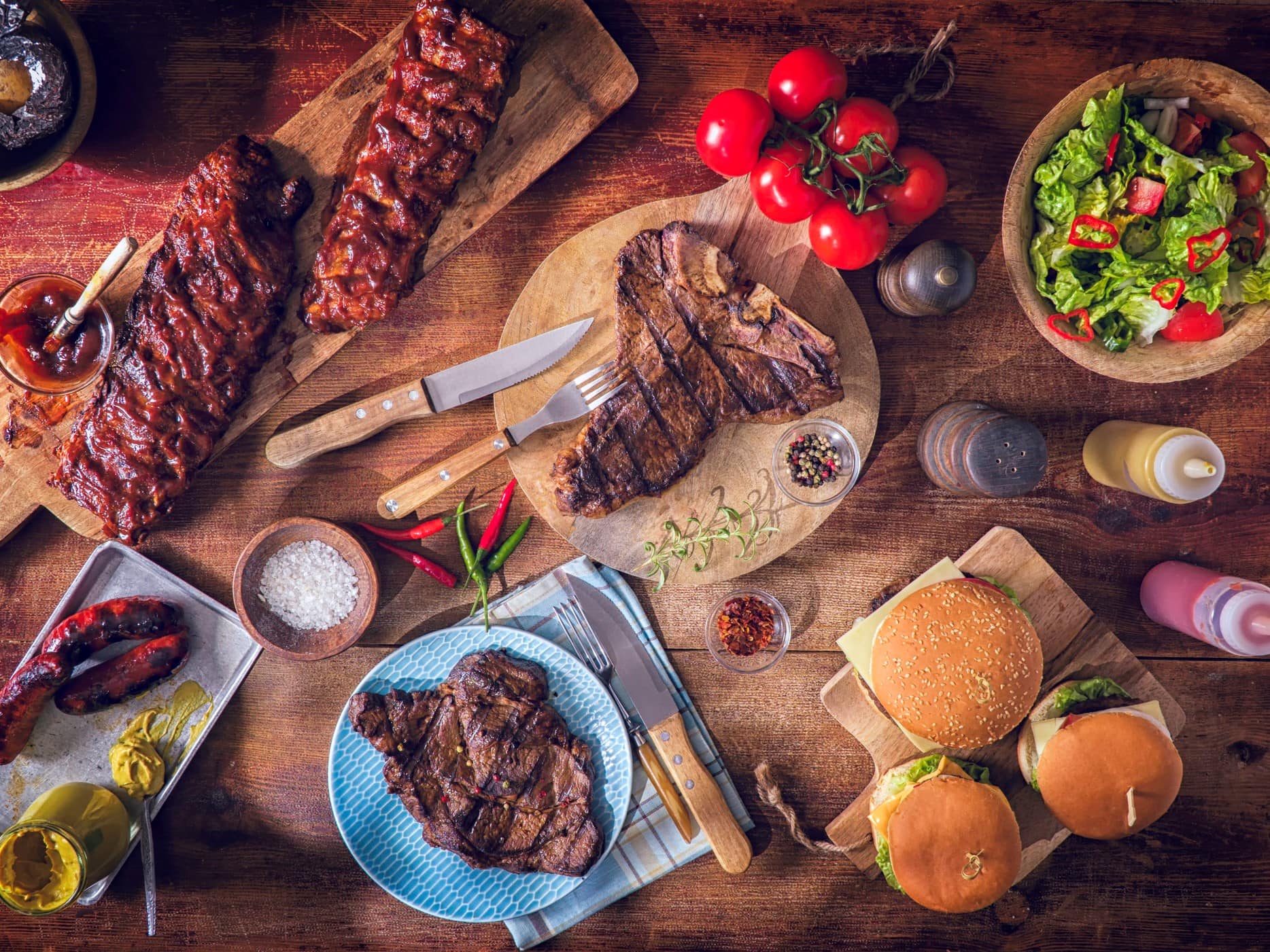 Cow-Sharing Produkte gegrillt auf Holzplatten serviert. Steaks, Würste, Burger und Rippchen. Dazu Salat und Gemüse als Dekoration. Aufnahme aus der Vogelperspektive.