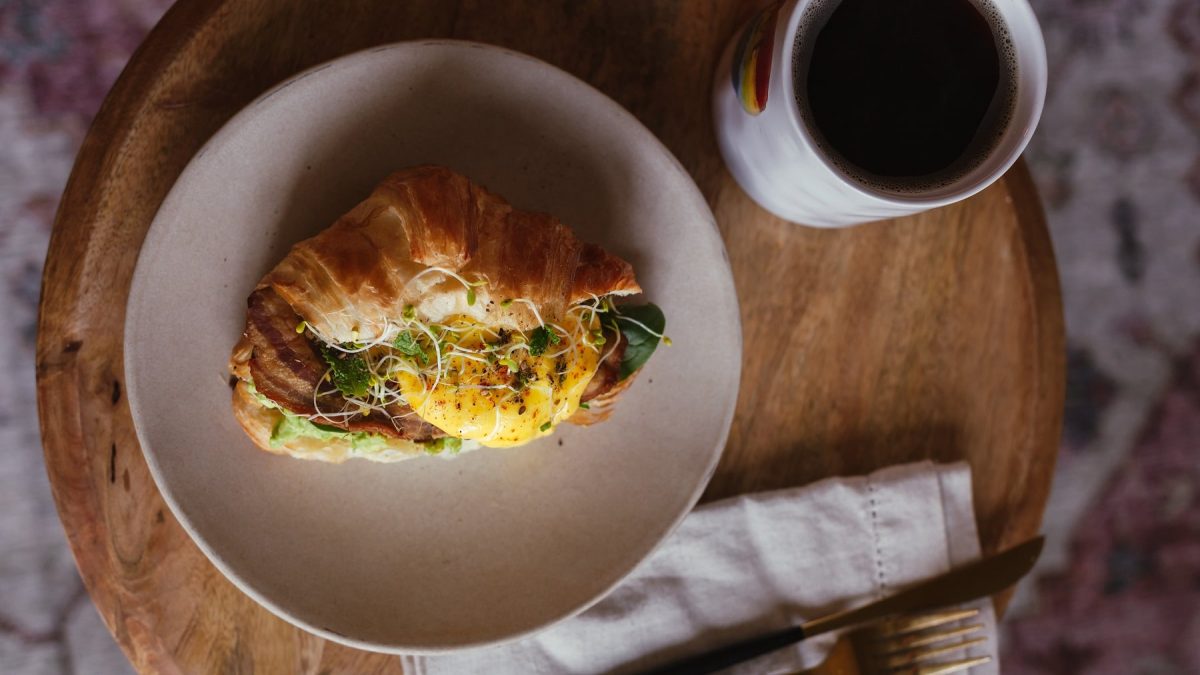 Ein belegtes Croissant mit Ei und Bacon auf einem beigen Teller. Daneben eine Tasse Kaffee und Besteck. Das Ganze auf einem kleinen Holztisch.