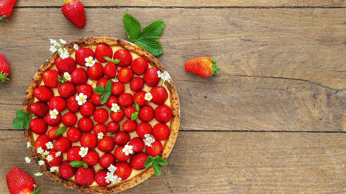 Auf einem Untergrund aus Holz steht ein runder Erdbeer-Vanille-Kuchen mit kandierten Blüten. Um ihn herum liegen Erdbeeren und Erdbeerblätter.