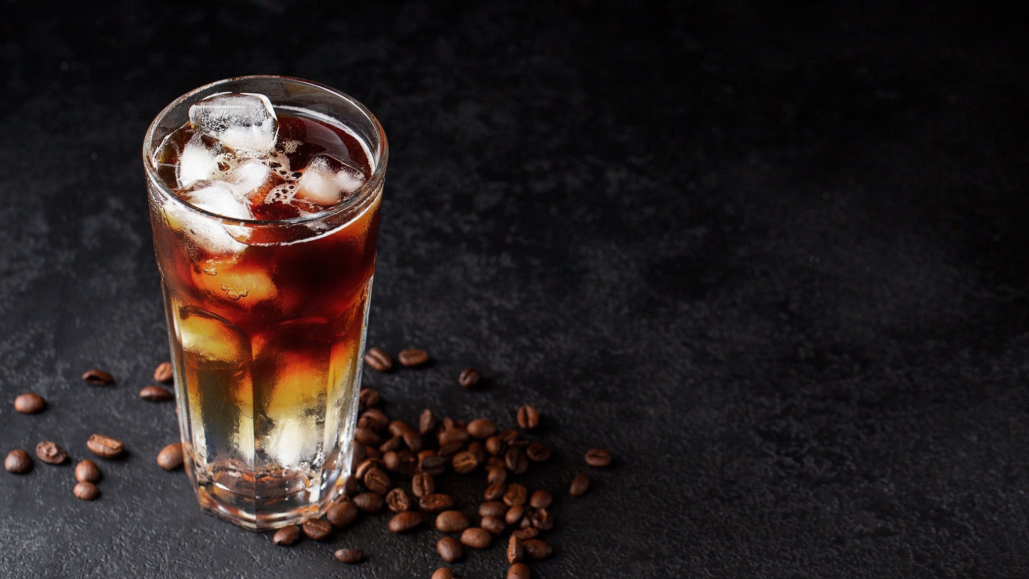 Ein Glas Espresso Tonic mit Eiswürfeln in dunklem Setting, daneben liegen Kaffeebohnen.