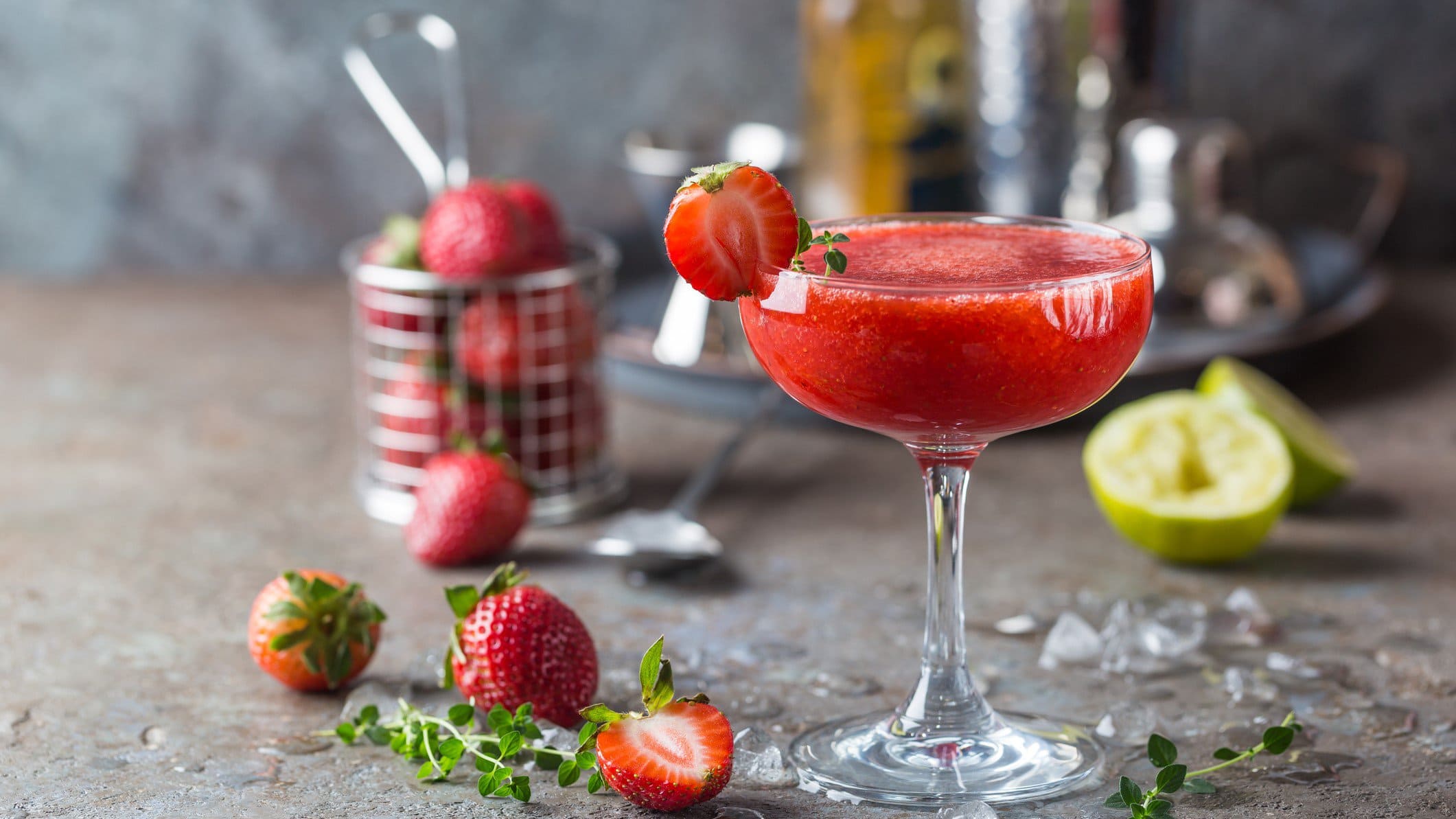 Frozen Strawberry Margarita in Martini-Glas auf braunem Untergrund vor grauem Hintergrund. Dahinter unscharf Erdbeeren im Metallkorb und Cocktail-Zubehör. Auf dem Untergrund Limetten, Erdbeeren und Thymian verteilt. Frontalansicht.