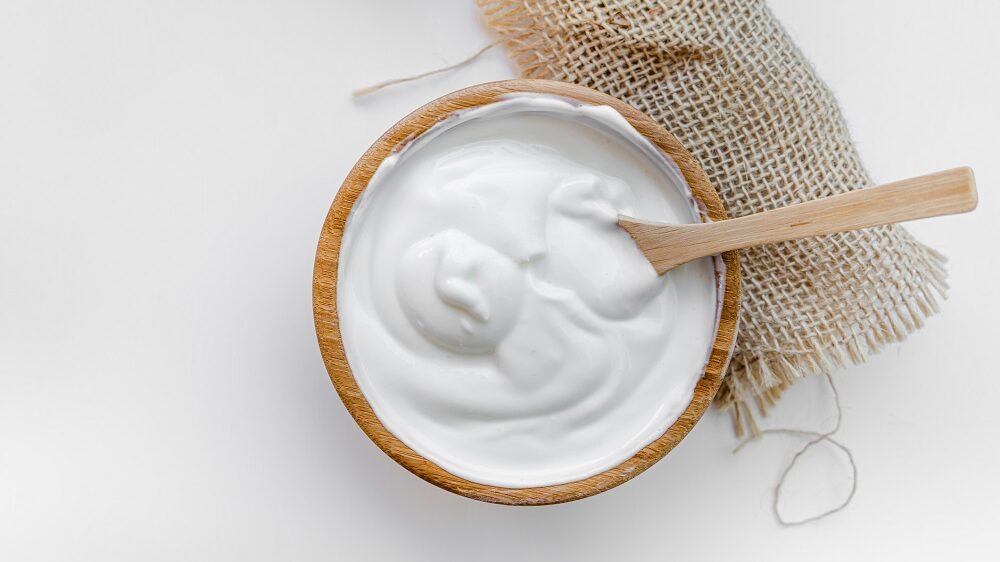 Aus der Vogelperspektive. In einer Schüssel aus Holz ist einen weiße Creme. Es handelt sich um selbstgemachten Joghurt. In dem Joghurt steckt ein Löffel aus Holz.