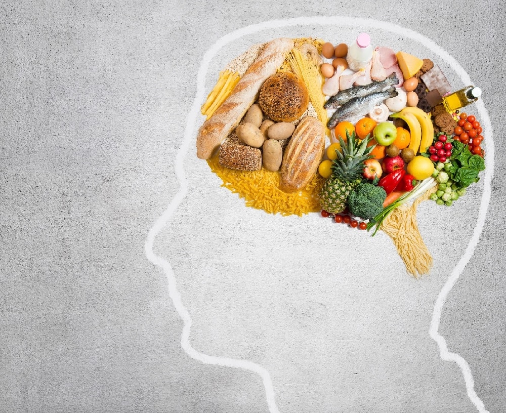Die Silhouette eines Kopfes mit einer weißen Linie auf grauem Untergrund. Im Bereich des Gehirns sind etliche Lebensmittel zu sehen.