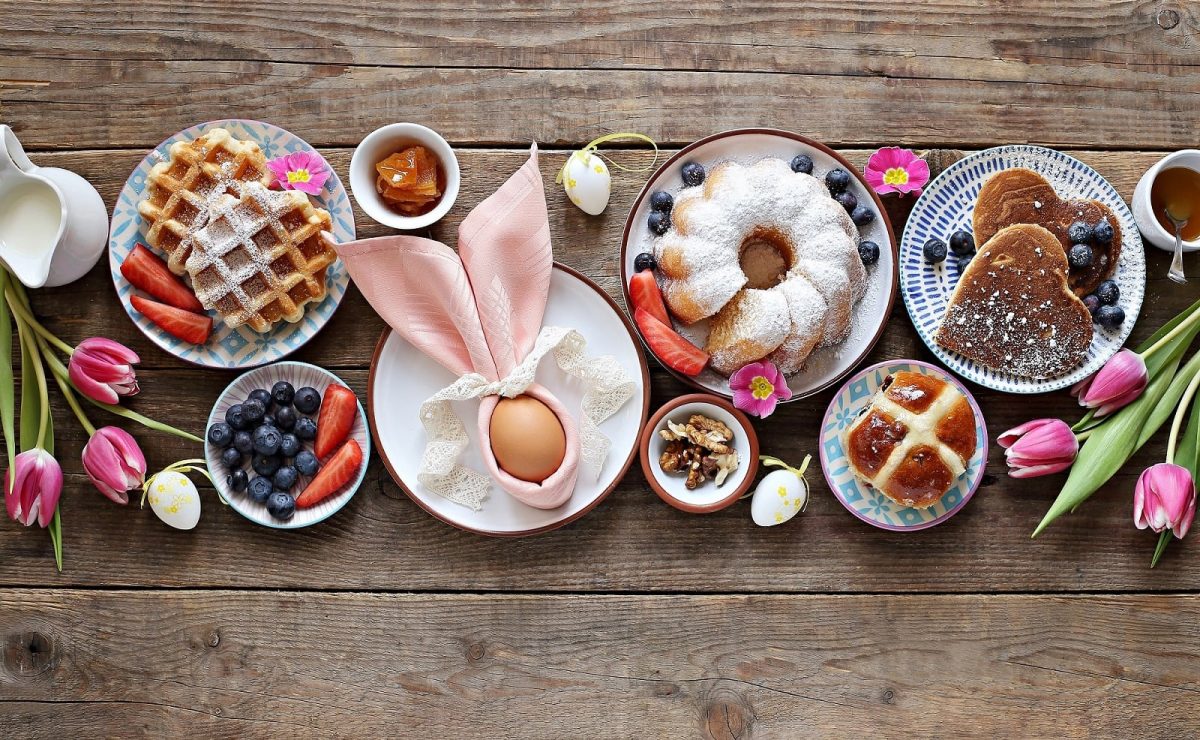 Ein Holztisch mit verschiedenen Osterbrunch-Rezepten beziehungsweise Gerichten wie Topfkuchen, Brötchen, Waffeln, und einem Ei.