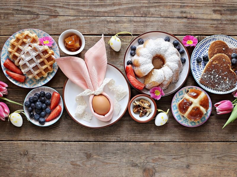 Ein Holztisch mit verschiedenen Osterbrunchrezepten beziehungsweise Gerichten wie Topfkuchen, Brötchen, Waffeln, und einem Ei.
