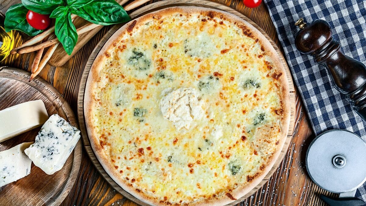 Auf einem Brettchen aus Holz liegt eine Gorgonzola-Pizza mit Rucola. Drumherum liegen Gorgonzola, TOmates, eine Pfeffermühl, Grisini und Basilikumblätter.