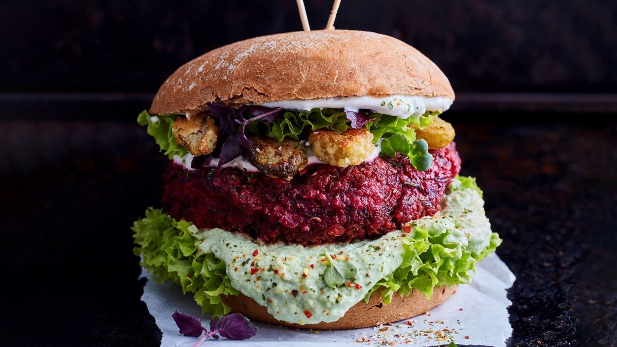 Ein Burger mit Namen "Purple Bliss Burger" mit rotem Bratling, Salat, weißer Kräutersauce und frittierten Gurken.