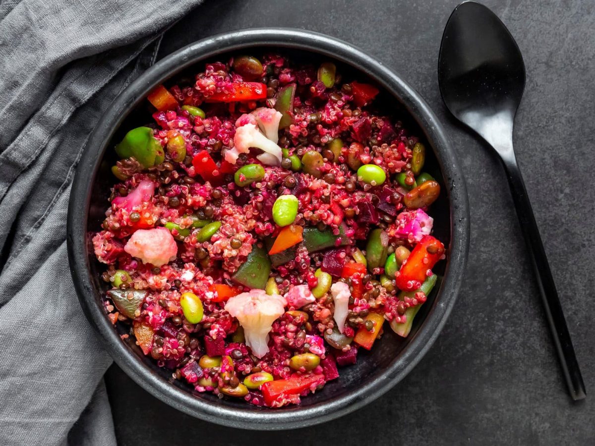 Eine schwarze Schale mit dem Rote Bete Quinoa und verschiedenem Gemüse auf einem dunklen Tisch mit grauer Tischdecke.