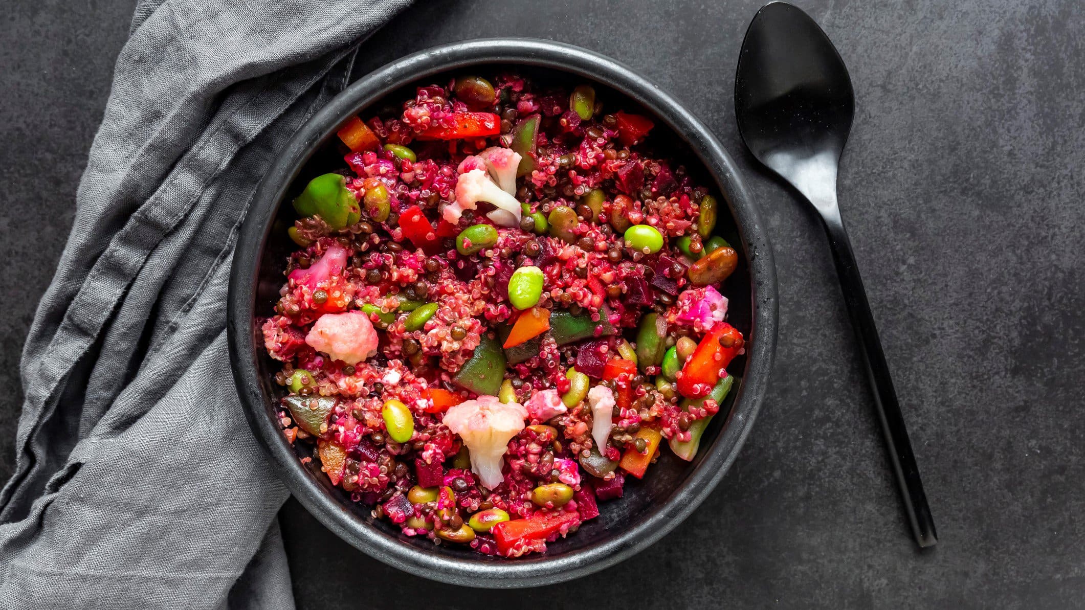 Eine schwarze Schale mit dem Rote Bete Quinoa und verschiedenem Gemüse auf einem dunklen Tisch mit grauer Tischdecke.