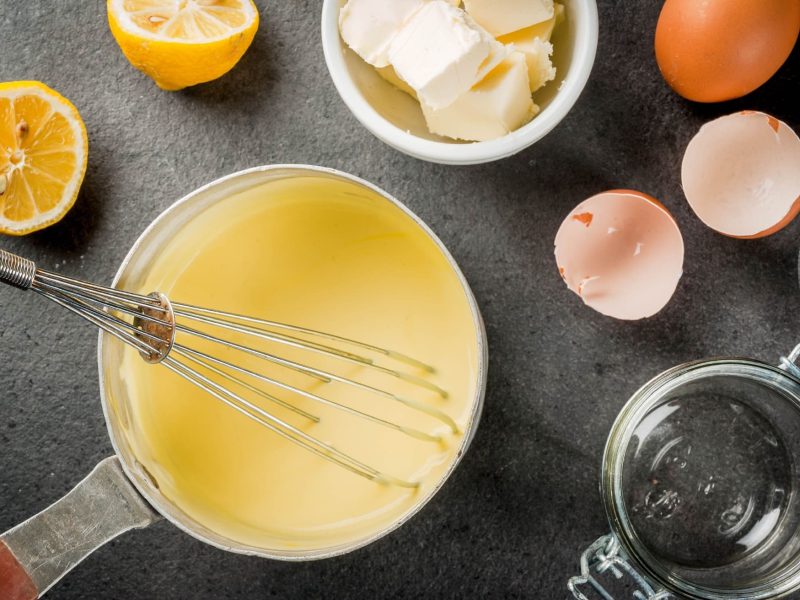 Auf einem Tisch aus Holz liegen Eierschalen, ausgepresste Zitronenhälften sowie Butterstückchen in einem Schälchen. In der Mitte steht ein gusseiserner Topf, in dem eine Sauce hollandaise ist. In der Sauce steht ein Schneebesen.