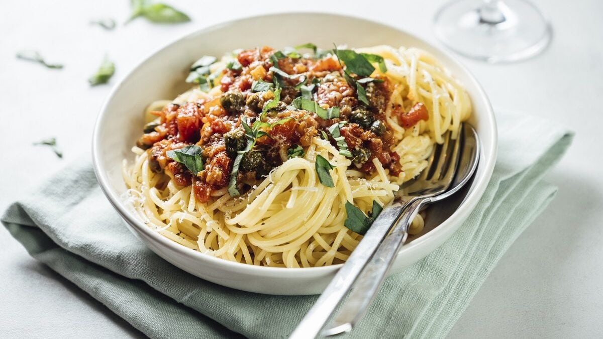 auf einem weißen Untergrund liegt ein graues, festes Tuch. Auf diesem steht ein weißer Teller. In dem Telle sind Spaghetti mit Tomaten-Sardellen-Sauce. In dem Teller liegen ein Löffel und eine Gabel.