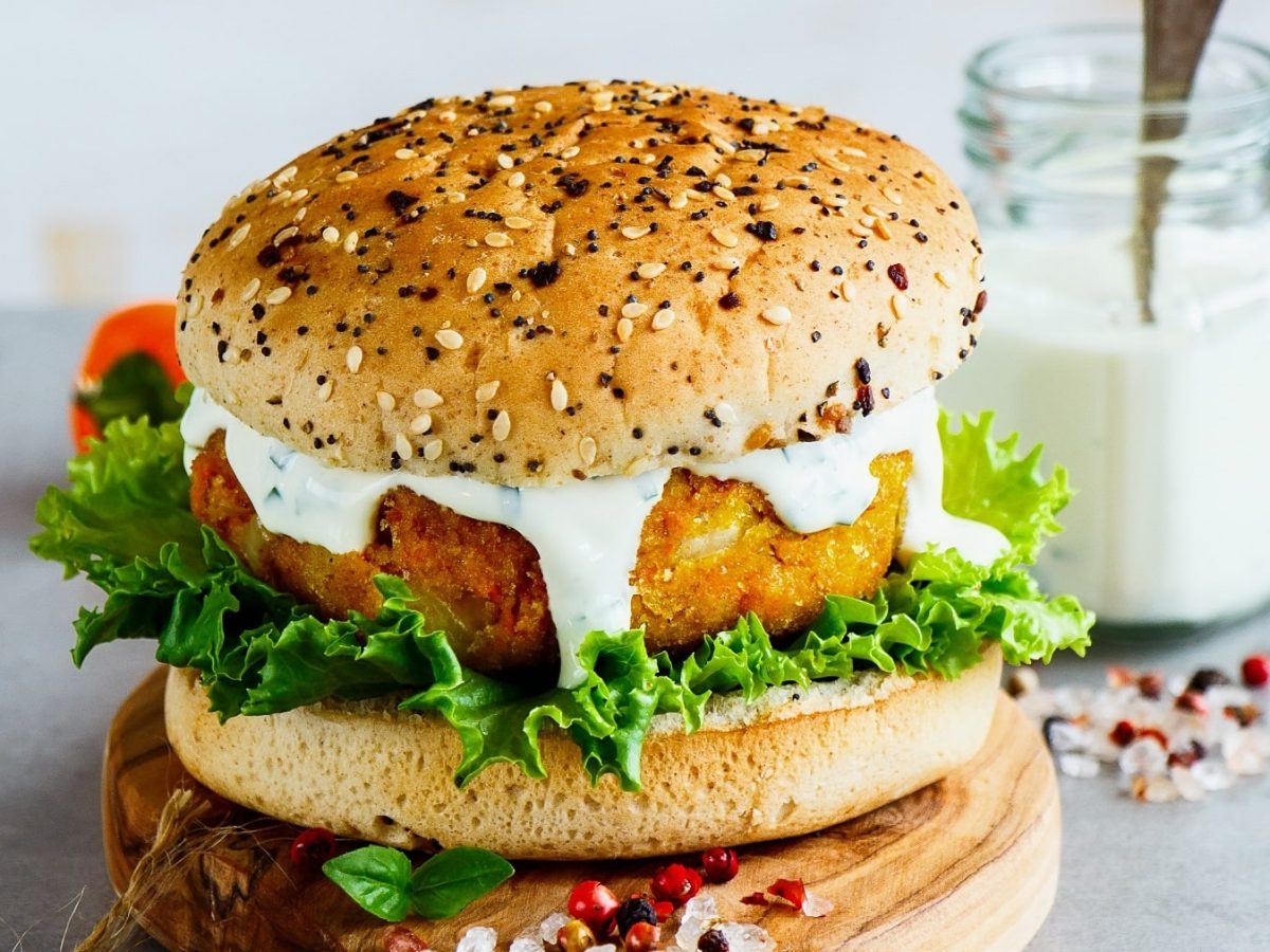 Ein veganer Burger mit Möhren-Hafer-Patty, Salat und weißer Sauce auf einem Holzbrett, dahinter steht ein Glas mit Mayo.
