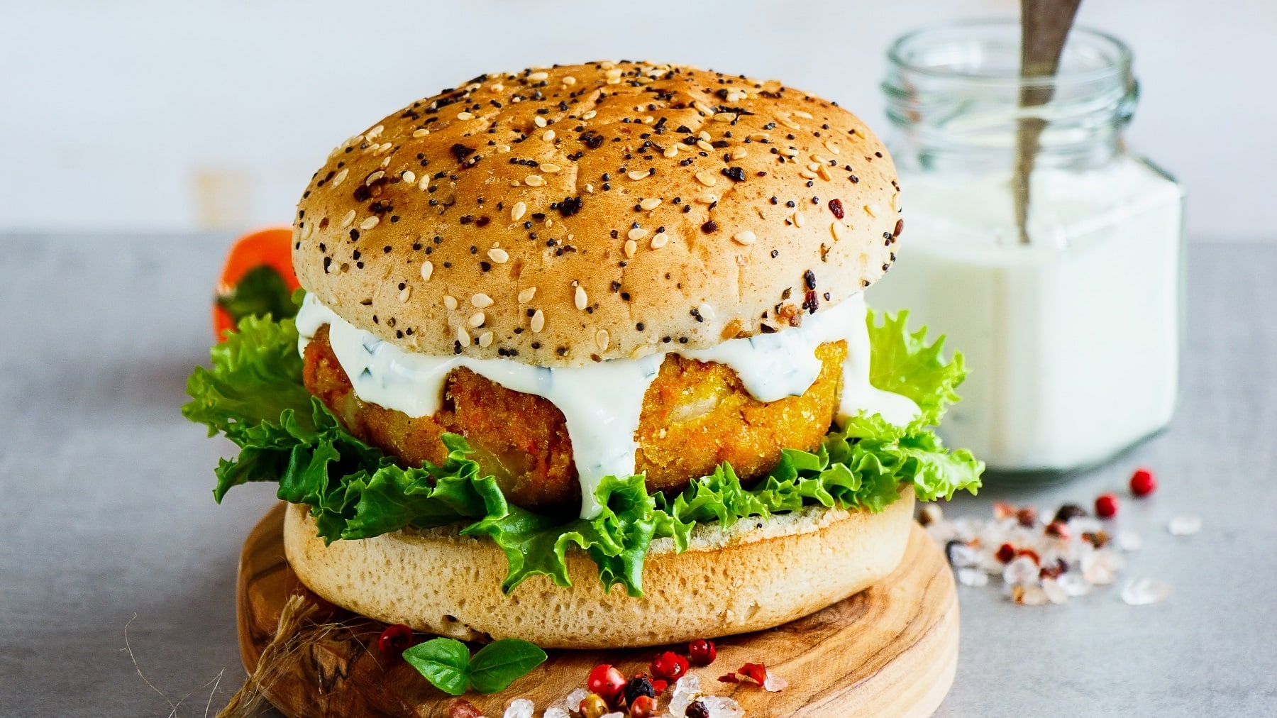Ein veganer Burger mit Möhren-Hafer-Patty, Salat und weißer Sauce auf einem Holzbrett, dahinter steht ein Glas mit Mayo.