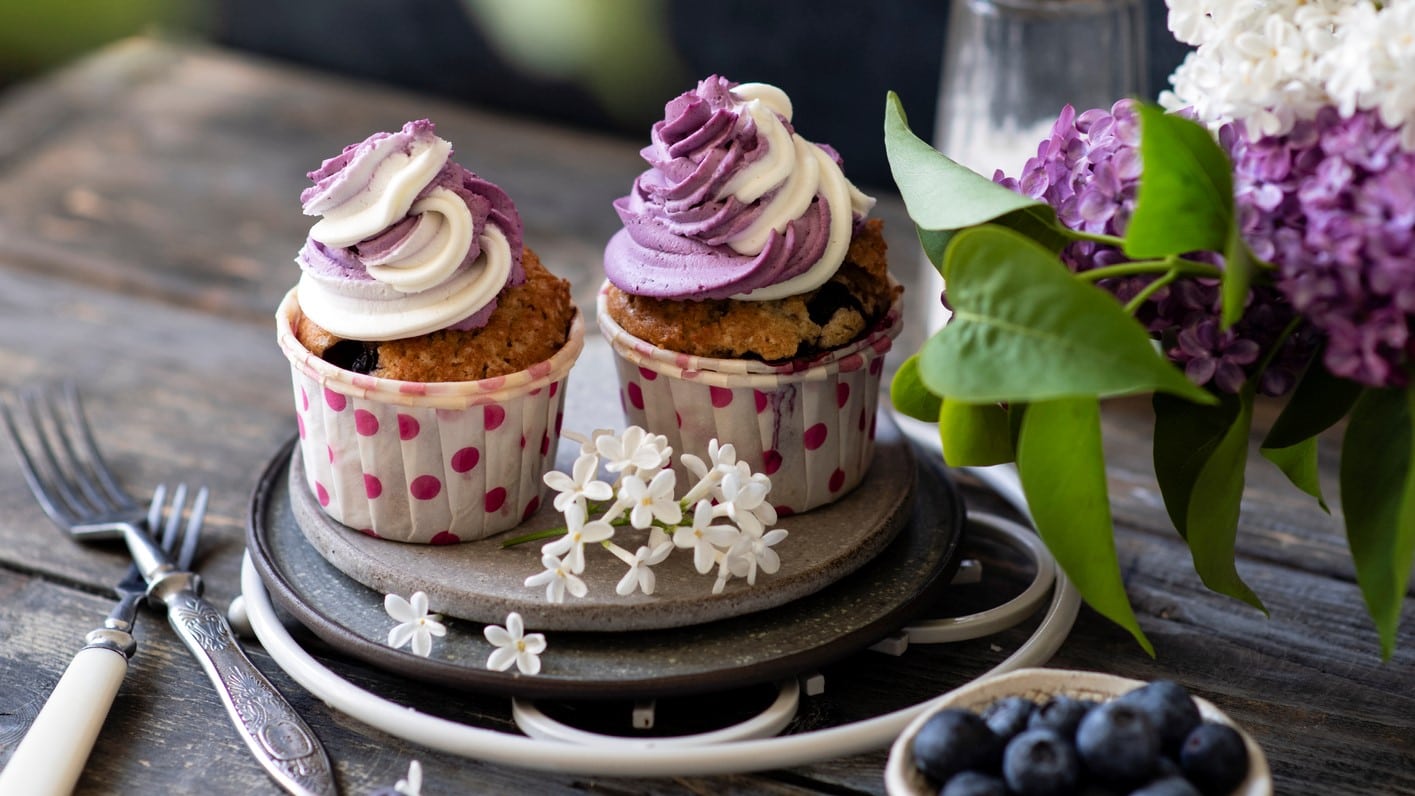 Zwei vegane Heidelbeer-Cupcakes auf einem Teller, daneben stehen frische Blumen und eine Schale mit Blaubeeren.