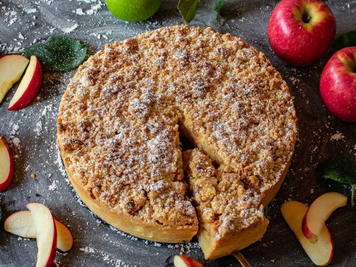 Auf einem Kuchenteller liegt ein veganer Apfel-Marzipan-Kuchen. Er ist mit Streuseln und Puderzucker bedeckt.