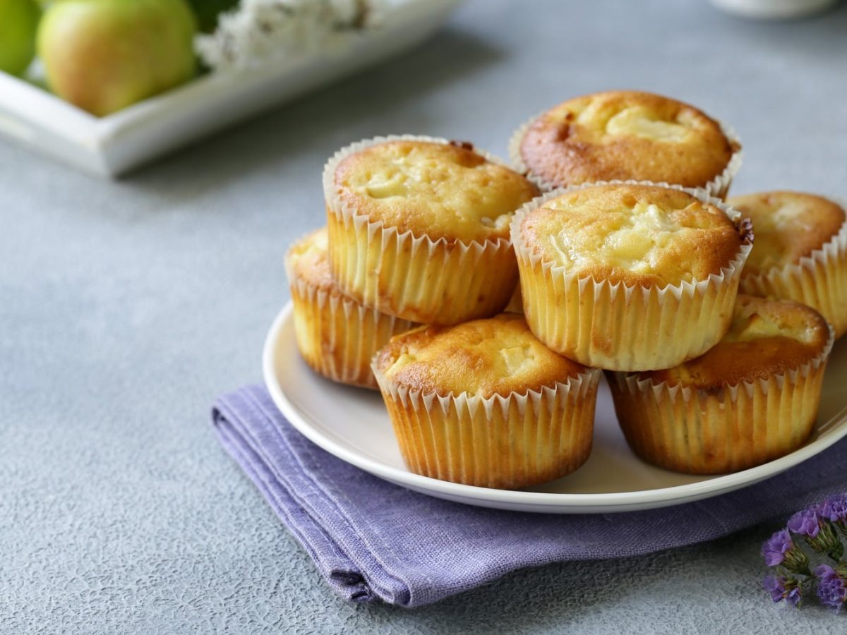 Mehrere Apfel-Muffins drapiert auf einem Teller. Angerichtet auf einem hellblauen Untergrund.
