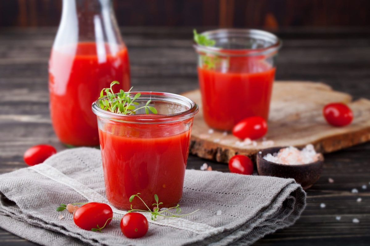 Zwei Gläser Tomatensaft und eine Karaffe mit frischen Tomaten auf einem grauen Geschirrtuch und einem Holzbrett