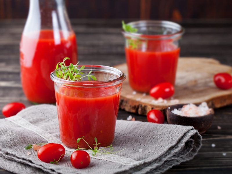 Zwei Gläser Tomatensaft und eine Karaffe mit frischen Tomaten auf einem grauen Geschirrtuch und einem Holzbrett