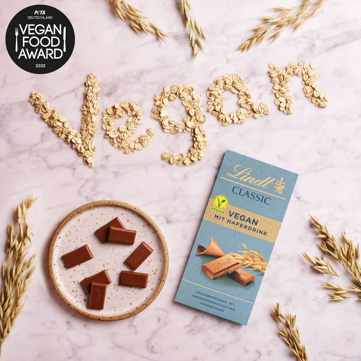Die Lindt Vegan Classic Schokolade, die den Vegan Food Award von PETA gewonnen hat, in der Draufsicht, daneben das Wort "Vegan" mit Haferflocken gelegt, mit Logo.