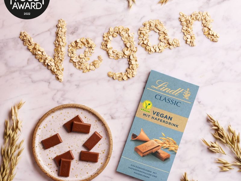 Die Lindt Vegan Classic Schokolade, die den Vegan Food Award von PETA gewonnen hat, in der Draufsicht, daneben das Wort "Vegan" mit Haferflocken gelegt, mit Logo.