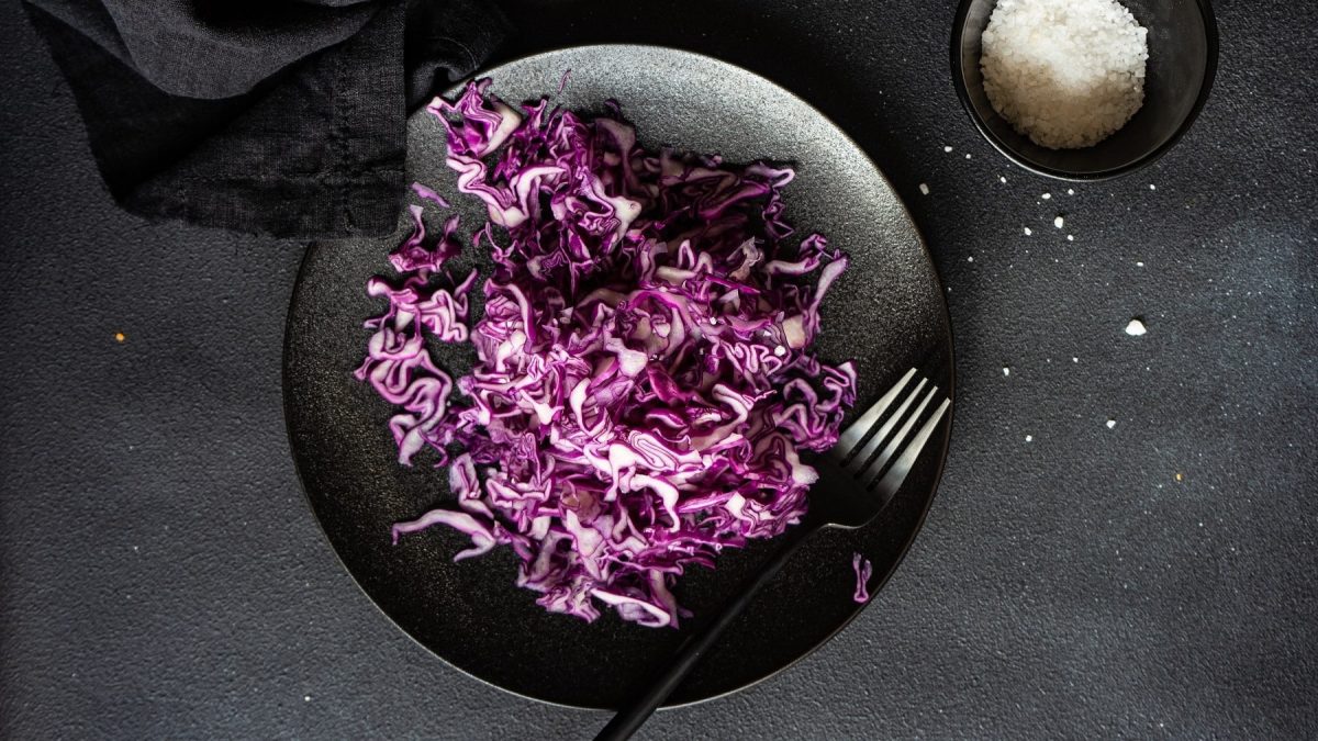 Rotkohl roh essen mit Salz bestreut und in dünne Streifen geschnitten, serviert auf einem schwarzen Teller auf dunklem Untergrund.