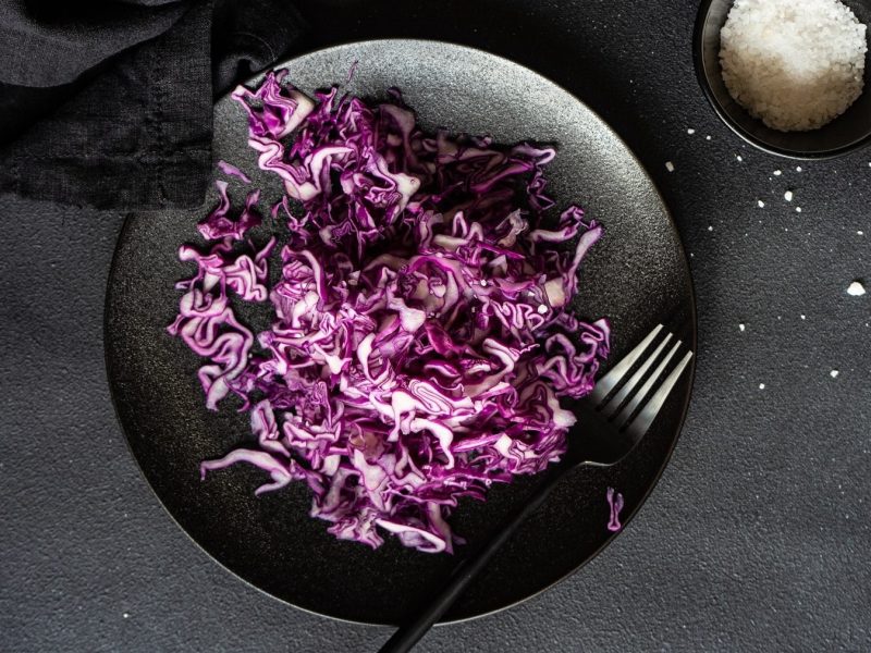 Rotkohl roh essen mit Salz bestreut und in dünne Streifen geschnitten, serviert auf einem schwarzen Teller auf dunklem Untergrund.