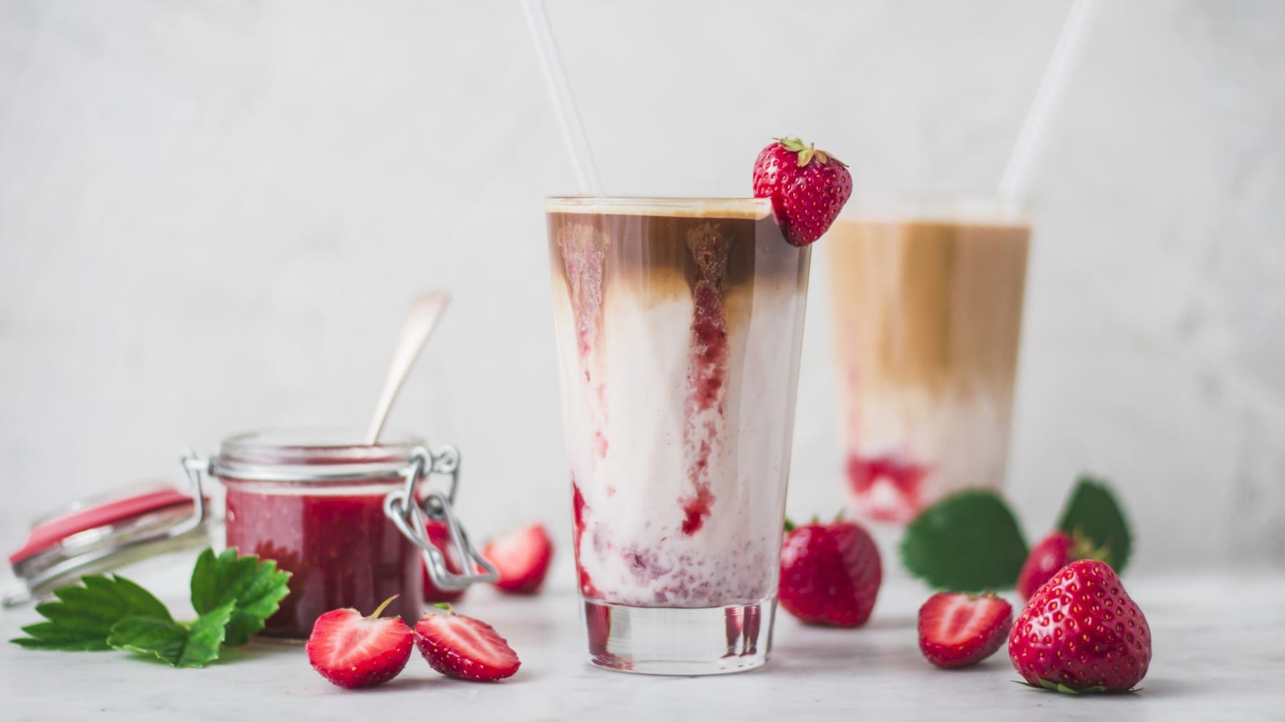 Ein Glas Iced Flat White mit Erdbeermilch und Erdbeersaice neben frischen Erdbeeren und einem Einmachglas