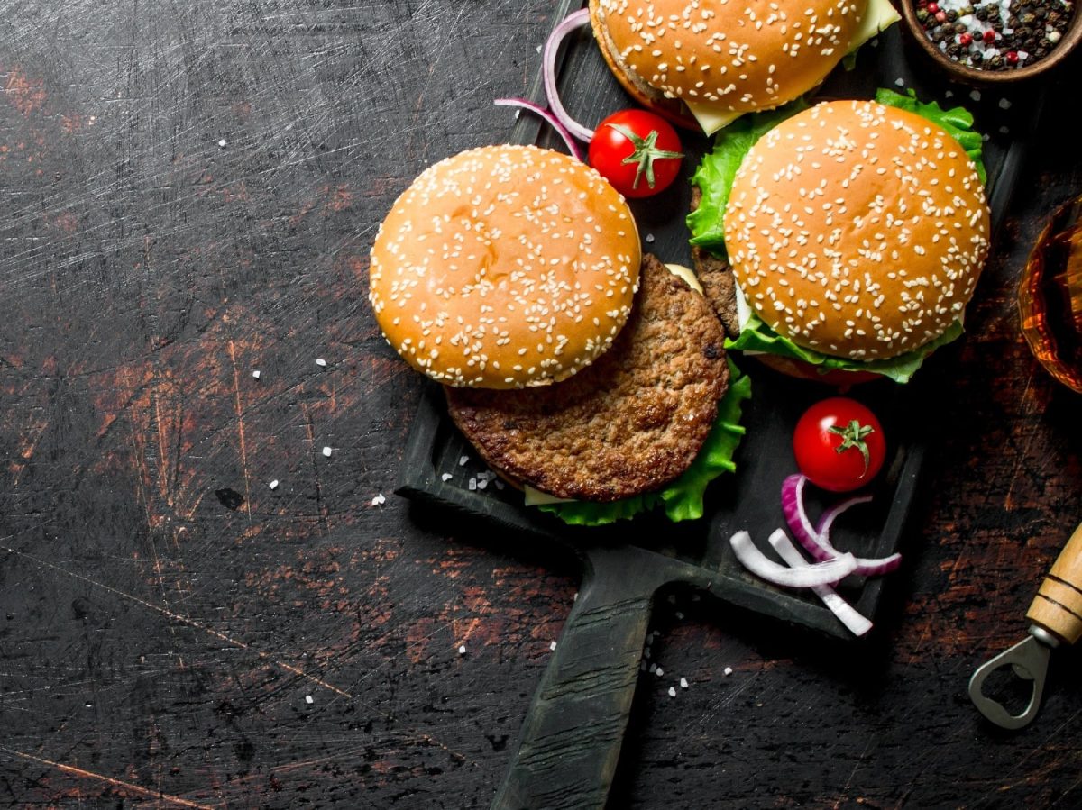 Drei Hamburger auf einem schwarzen Schneidebrett mit Tomaten, roten Zwiebeln und Pfeffer.