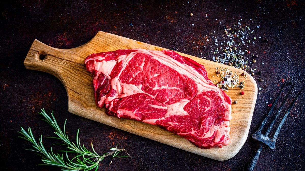 Ein rohes steak auf einem Holzbrett und schwarzem Untergrund. Umgeben von einer Gabel, Pfeffer und Kräutern.