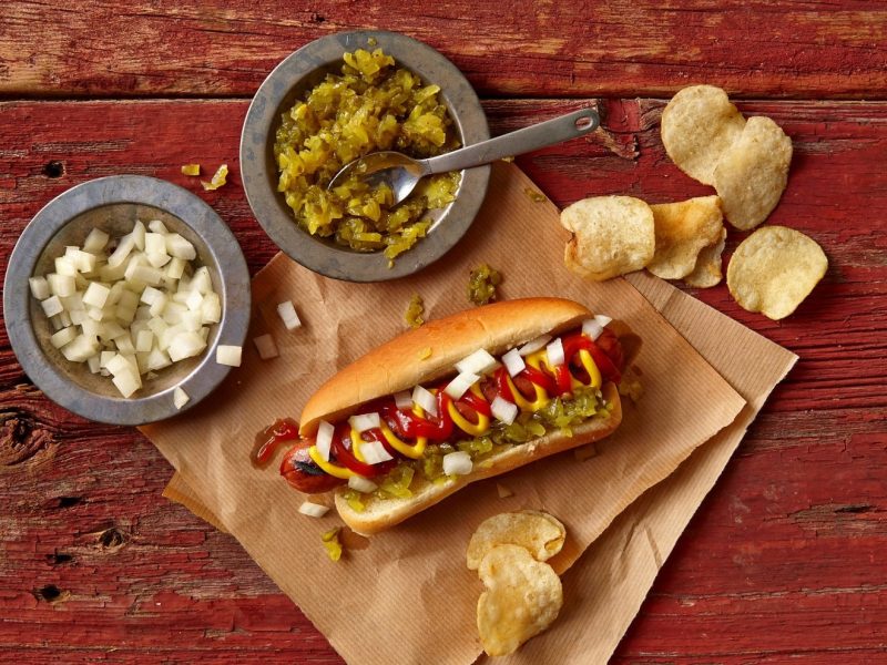 Woher kommt der Name Hotdog? Dazu ein Hotdog auf Backpapier mit Kartoffelchips. Daneben je eine graue Schale mit gewürfelten Zwiebeln und sauren Gurken auf rotem Holztisch. Aufnahme aus der Vogelperspektive.
