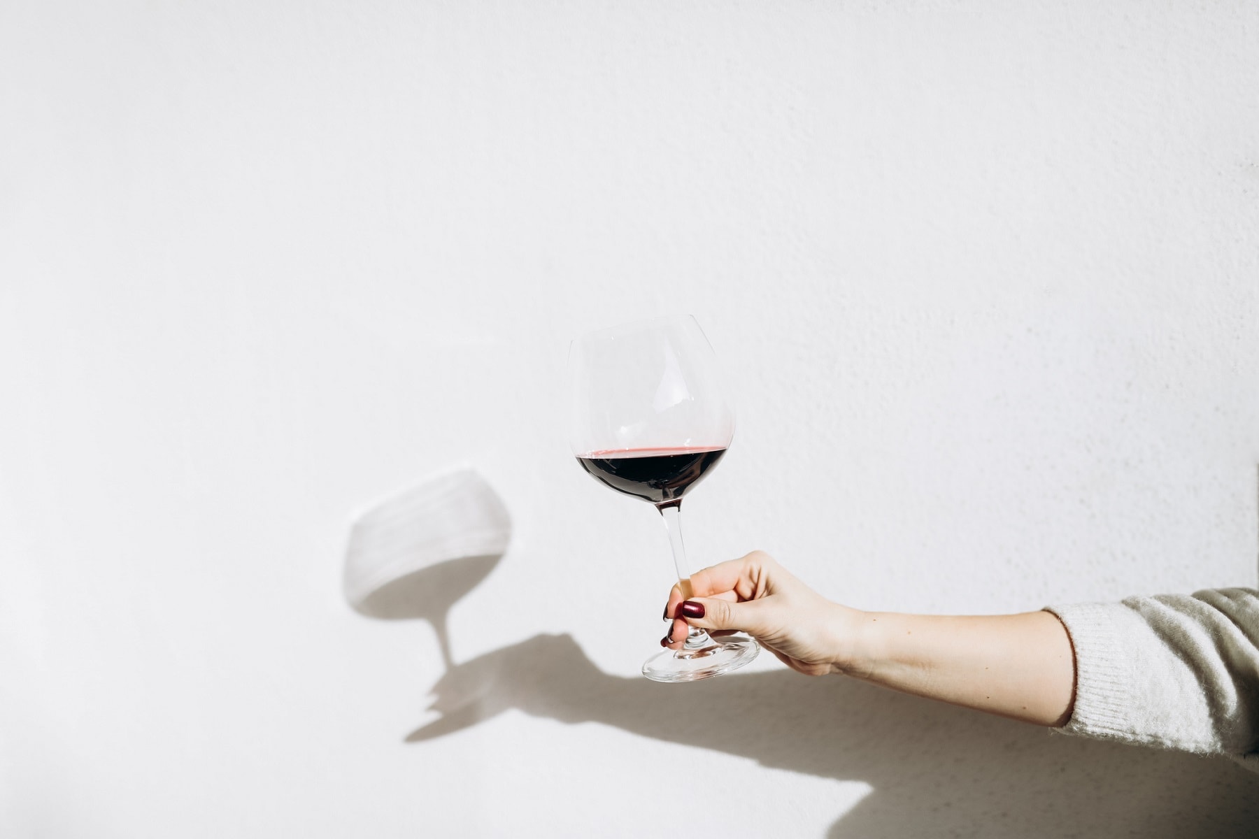 Ist Wein vegan? Eine Frau hält ein Glas Rotwein vor einer weißen Wand.
