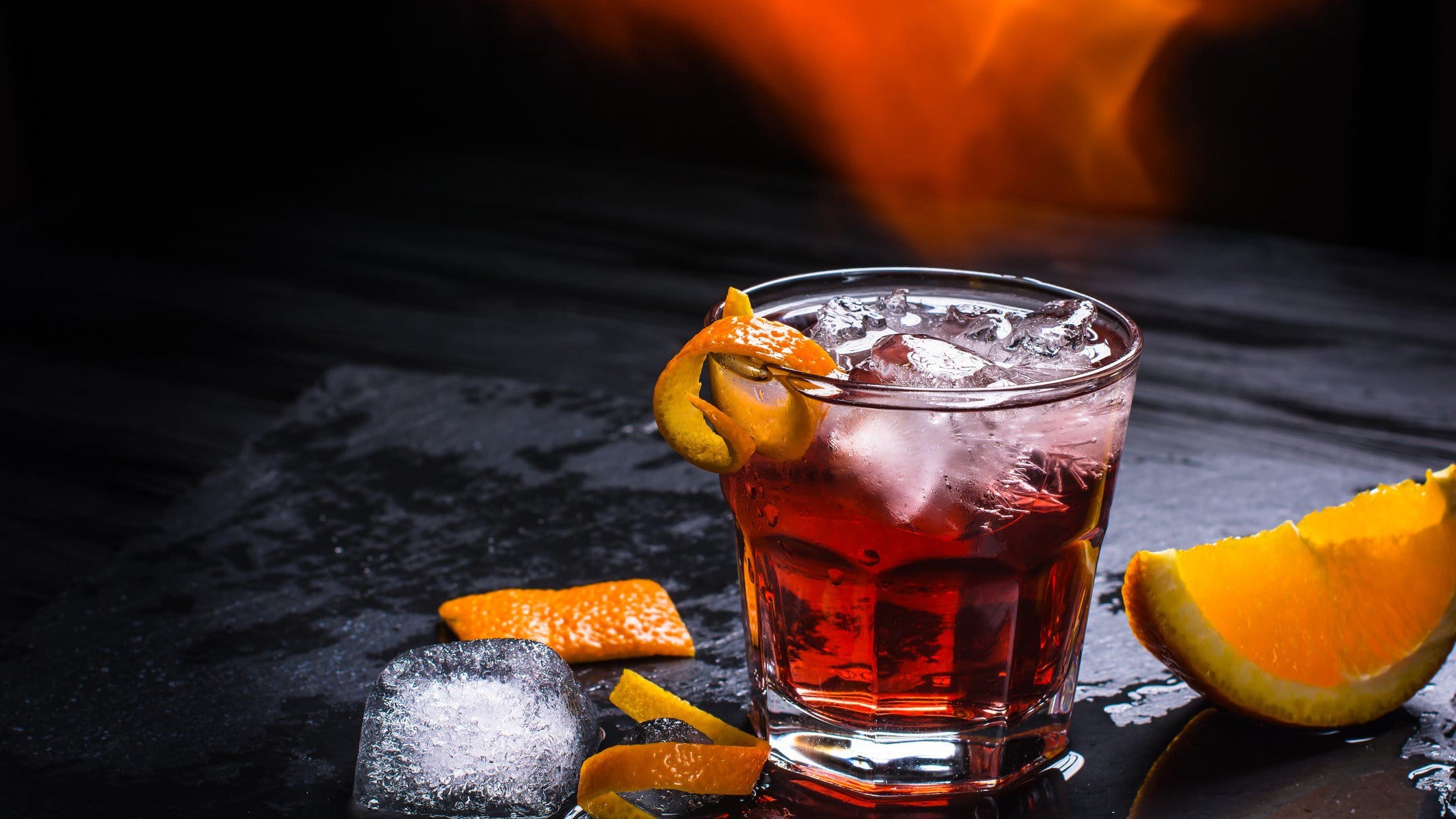 Mezzoni Cocktail mit Mezcal wie Negroni mit Eis und Orangenschale auf schwarzem Untergrund. Einswürfel und Orange als Dekoration im Bild. Flammen über dem Shortdrink. Frontalansicht.