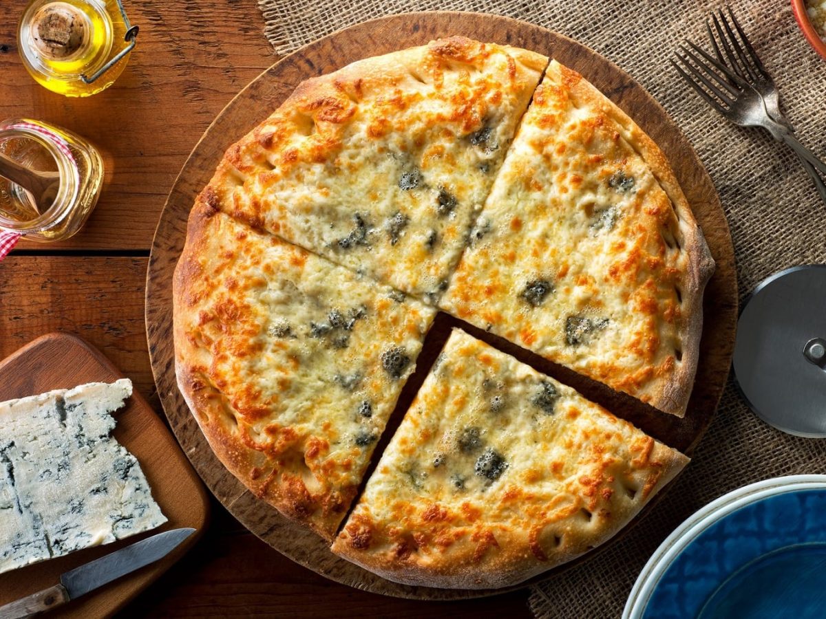 Pizza Gorgonzola mit Honig auf einem Holzbrett. Daneben stehen verschiedene Zutaten und Küchenutensilien. Das Ganze auf einem hölzernen Untergrund.
