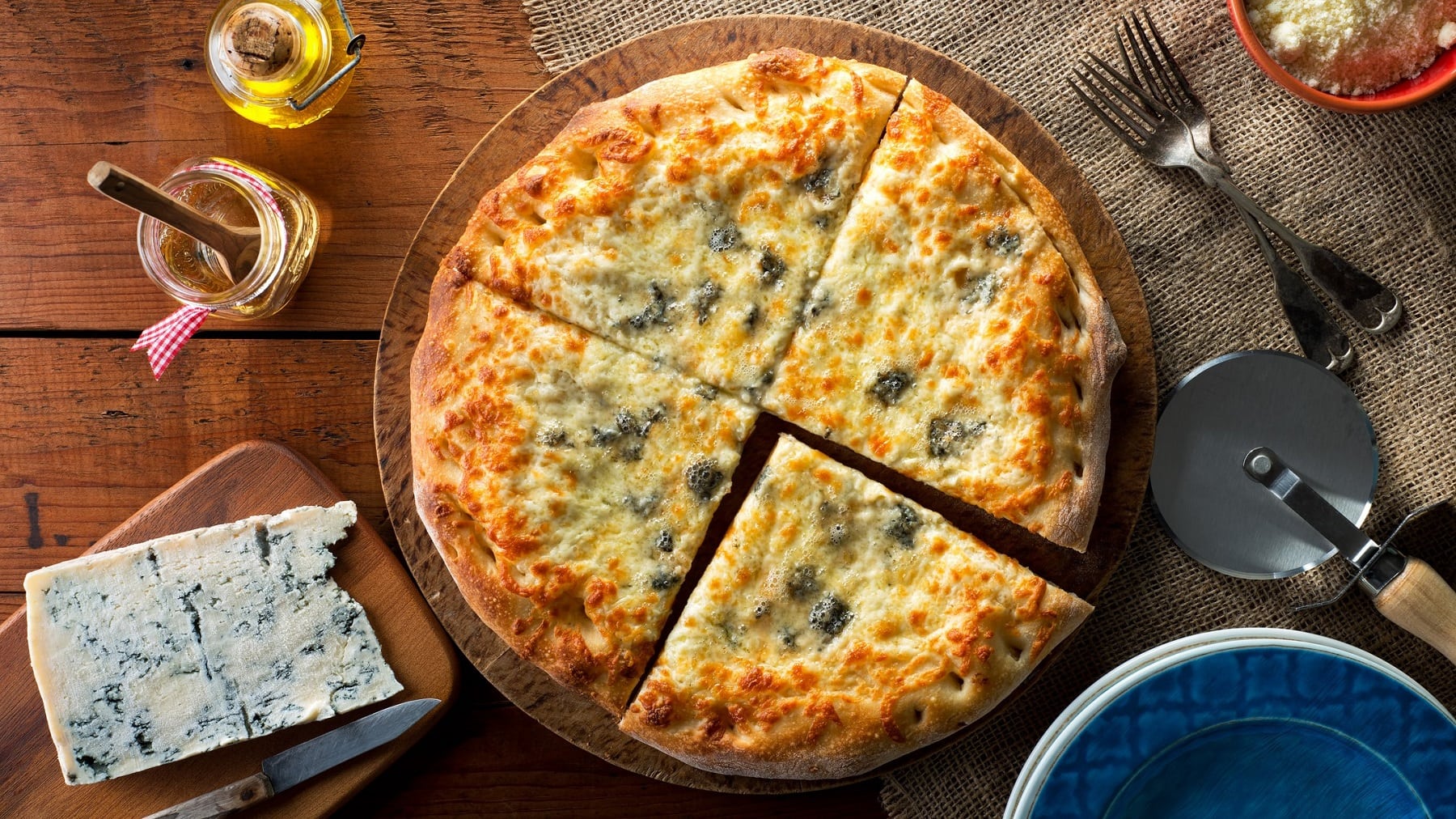Pizza Gorgonzola mit Honig auf einem Holzbrett. Daneben stehen verschiedene Zutaten und Küchenutensilien. Das Ganze auf einem hölzernen Untergrund.