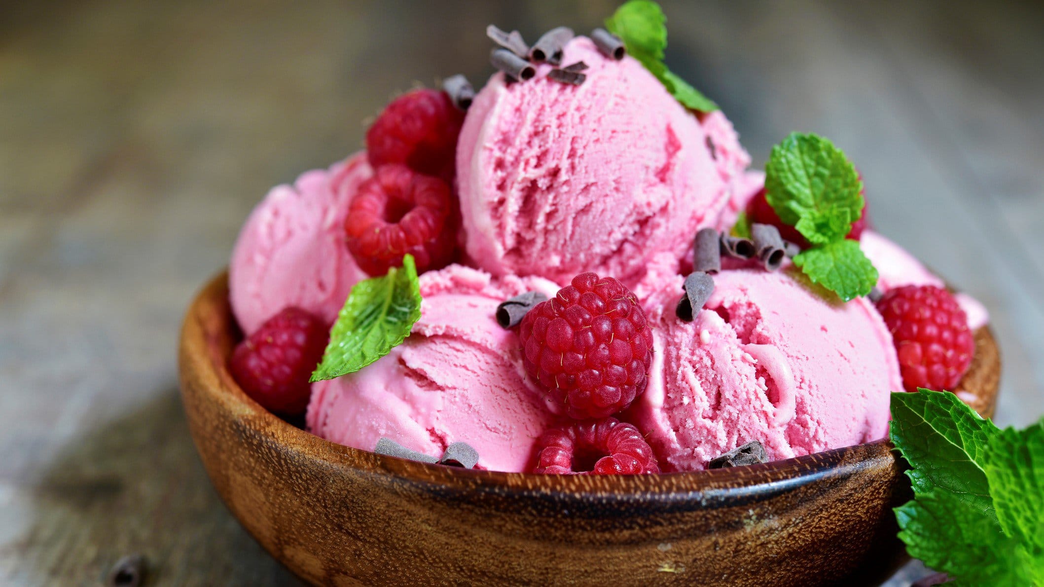 Blitzschnelles Himbeer-Joghurt-Eis mit frischen Himbeeren, Minze und Schokoladenflocken in Tonschale auf hellem Holztisch. Frontalansicht.