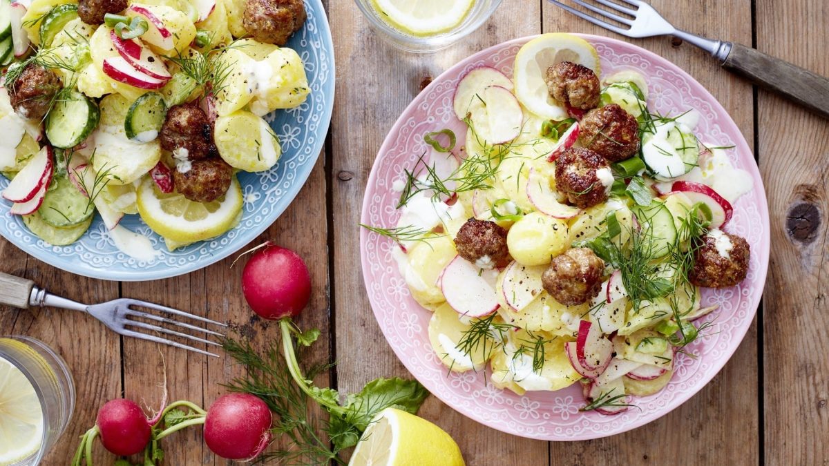 Skandinavischer Kartoffelsalat mit Frikadellen, Radieschen, Gurke, Dill und frischem Skyr, serviert auf rosanen Tellern auf einem Holztisch.