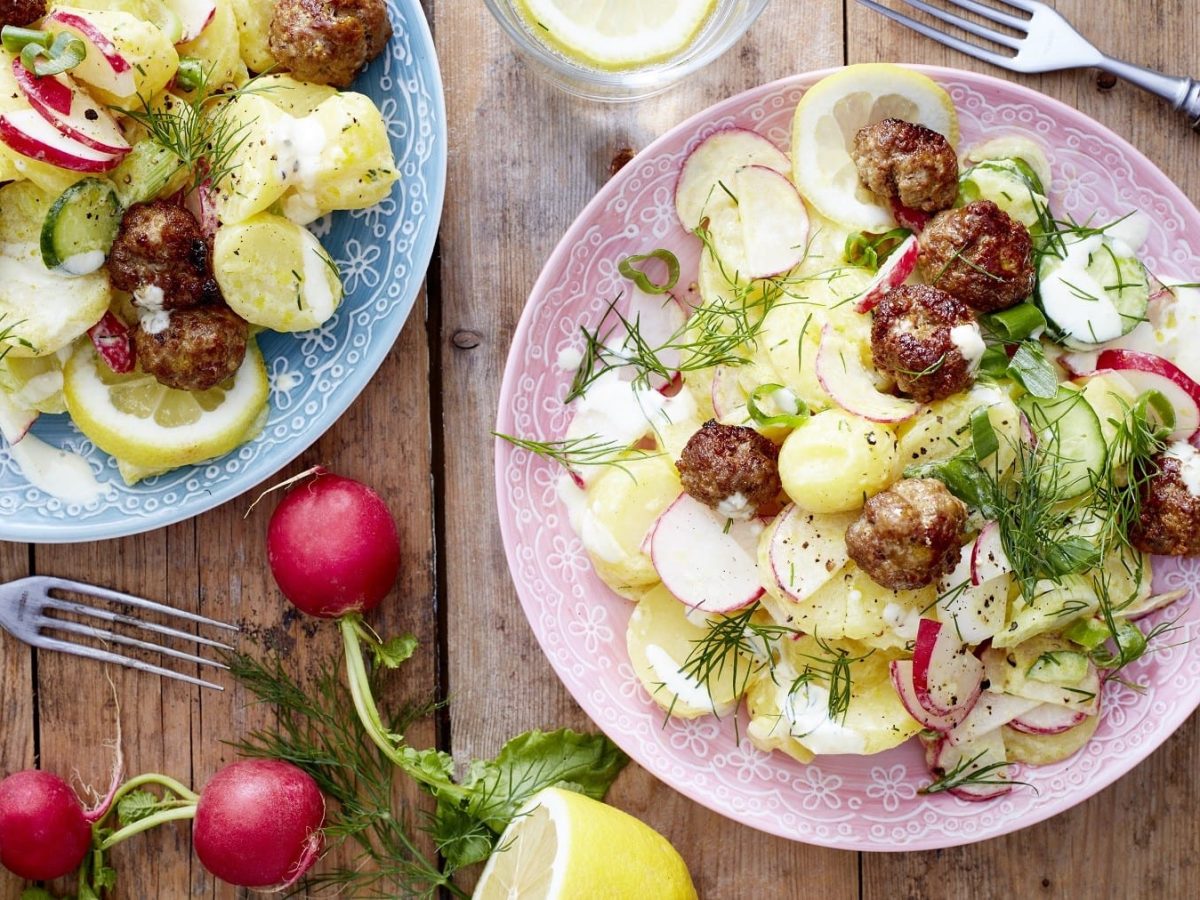 Skandinavischer Kartoffelsalat mit Frikadellen, Radieschen, Gurke, Dill und frischem Skyr, serviert auf rosanen Tellern auf einem Holztisch.