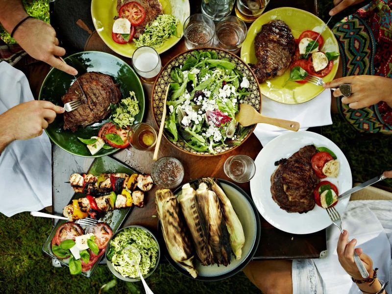 Tag des Barbecues verschieden Grillgerichte auf Tellern auf Tisch. Händ mit Messern und Gabeln schneiden Fleisch. Auhnahme aus der Vogelperspektive.
