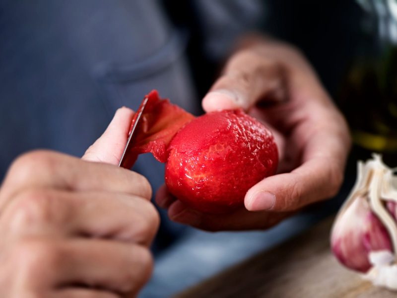 Ein Mensch häutet eine Tomate. Man sieht nur dessen Hände, die ein Messer und eine hast gehäutete Tomate halten.