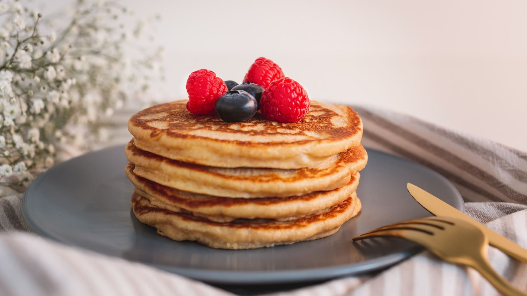 Ein Stapel vegane Pancakes mit frischen Beeren auf einem dunkelgrauen Teller, daneben goldenes Besteck.