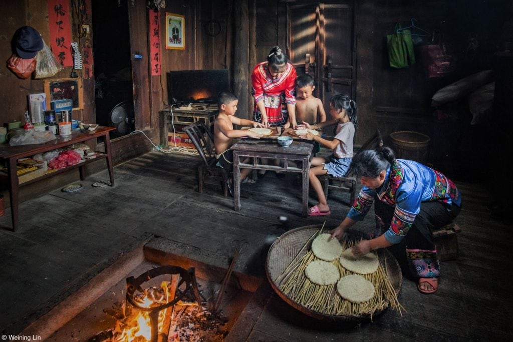 Eine asiatische Familie ist um einen Tisch versammelt, daneben bereitet eine der Frauen Teigfladen auf einem Ofen auf dem Boden zu.