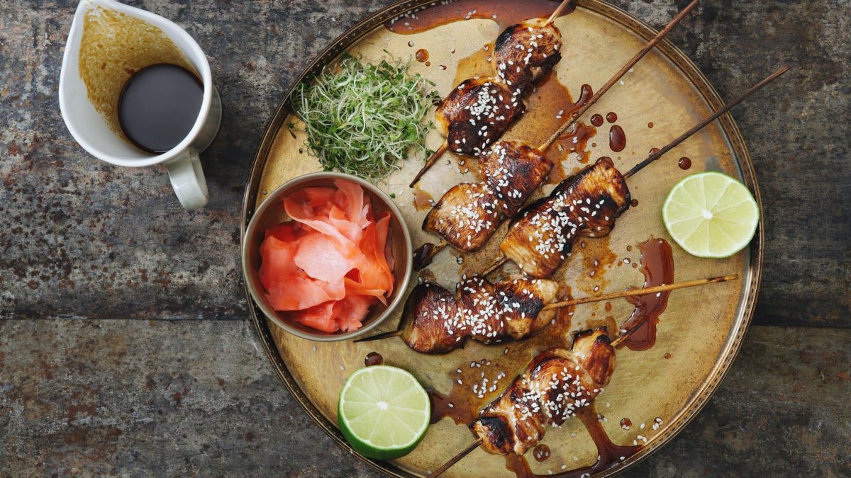 Gegrillte Yakitori japanische Hähnchenspieße mit Sesam, Limette, Kresse und einer Schale eingelegtem Ingwer auf beigem Tonteller. Daneben weiße Kanne mit Ponzu-Sauce auf dunklem Holz. Aufnahme aus der Vogelperspektive.