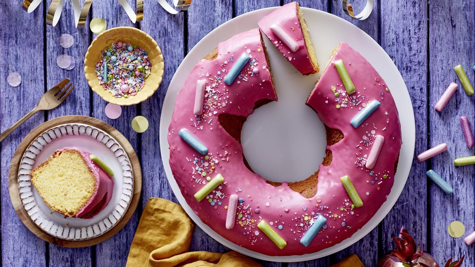 Auf einem lila Tisch steht ein Teller mit einem Donut-Kuchen. Er ist mit Streuseln und Kaubonbons verziert.