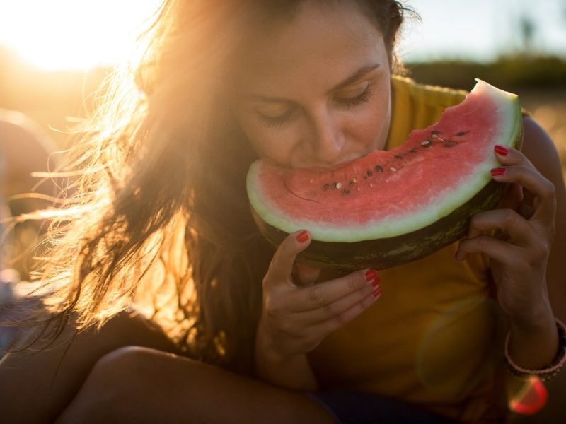 Eine junge Frau beißt beleuchtet von der Abendsonne in eine Scheibe Wassermelone.