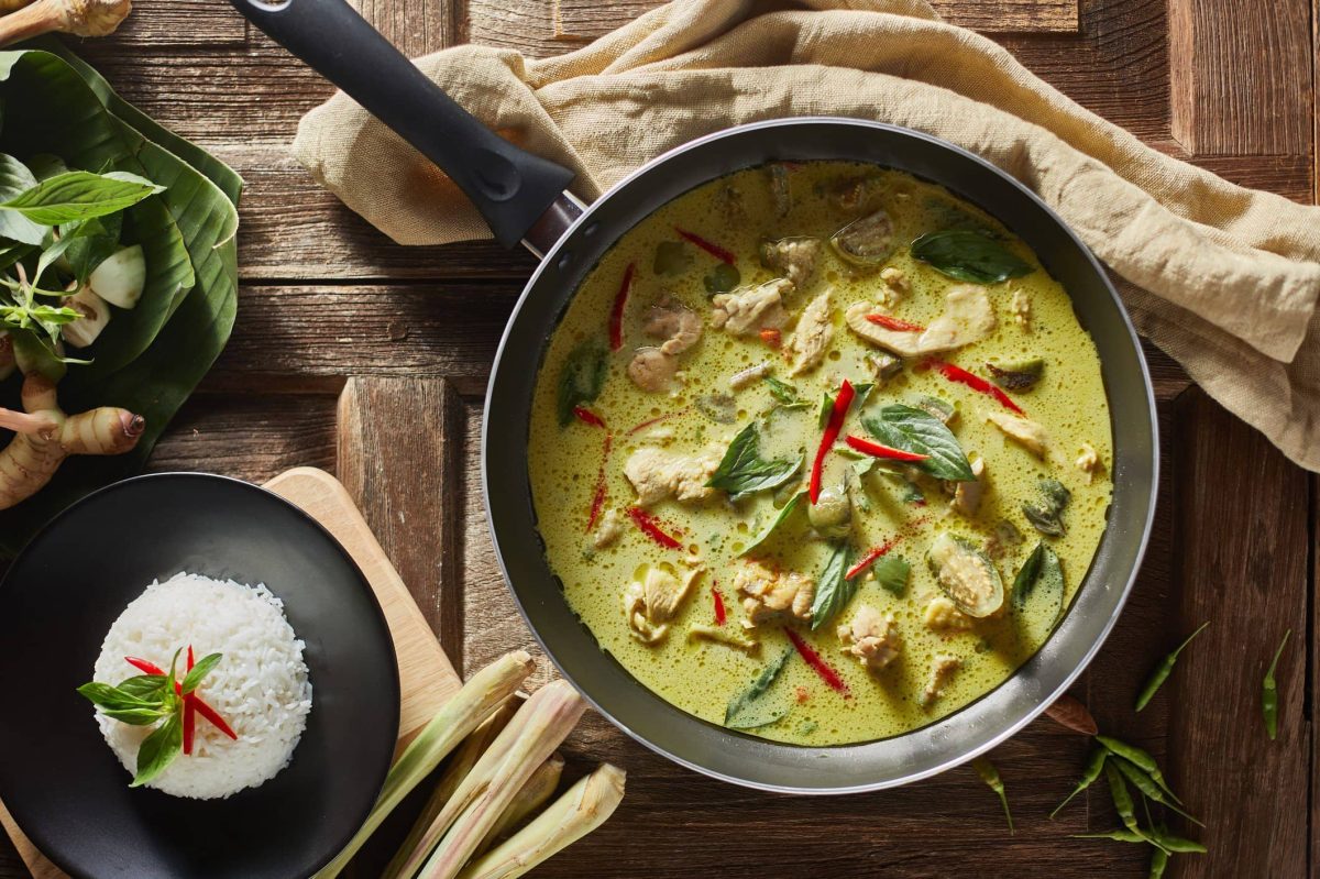 Eine Pfanne mit grünem Thai-Curry neben einer Schüssel Reis auf braunem Geschirrtuch