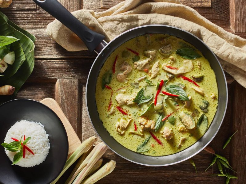 Eine Pfanne mit grünem Thai-Curry neben einer Schüssel Reis auf braunem Geschirrtuch