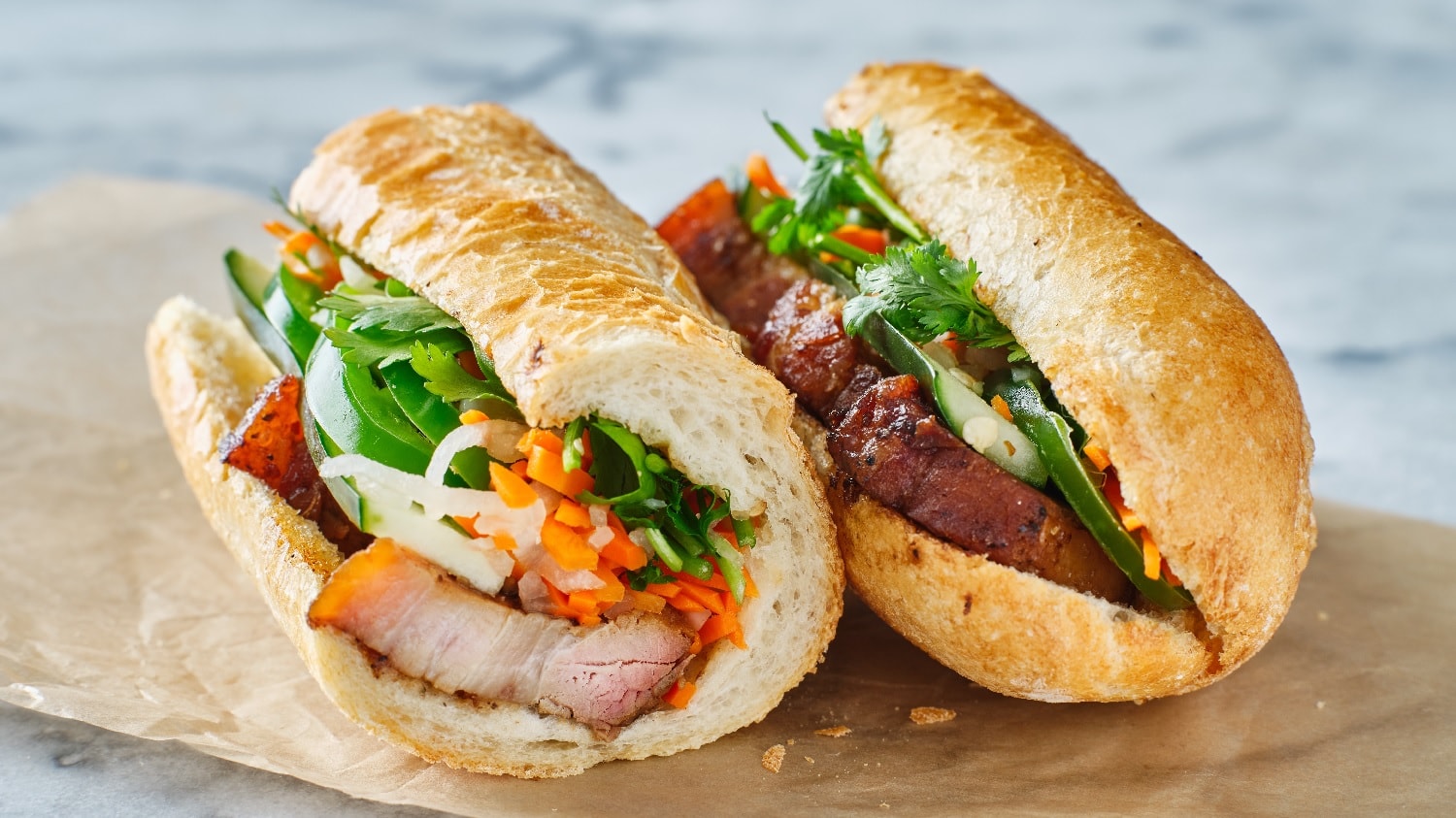 Zwei Banh-mi Sandwiches auf einem Marmortisch mit etwas Backpapier. Das Sandwich ist angeschnitten, sodass man den Inhalt (Karotten, Paprika, Schweinefleisch und Koriander) sehen kann.